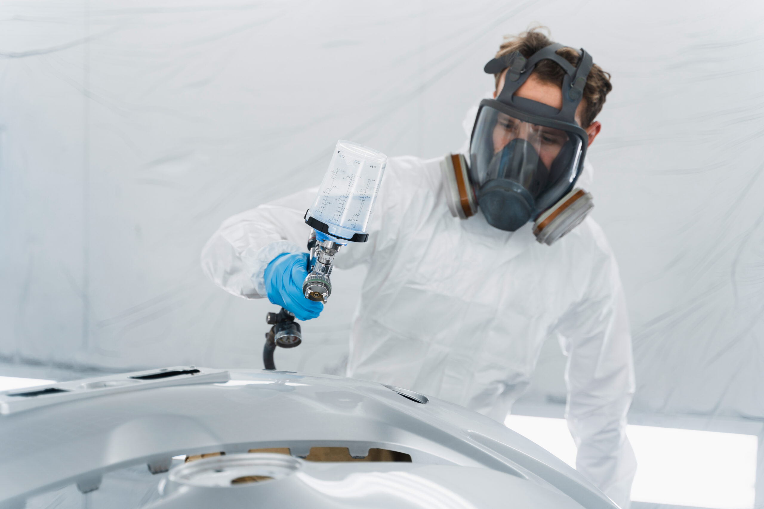 Car painter in a protective suit and mask paint a auto bumper in a painting booth. Vehicle body repair process Automotive mechanical engineer painting the body of a red car with special suit at Paint and Body Shops.